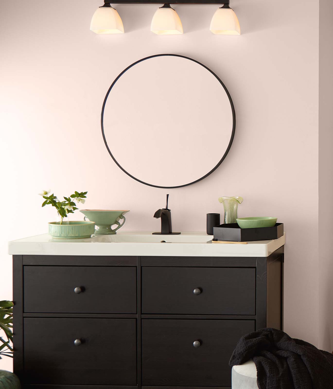 A bathroom with a white sink, brown cabinet and large round mirror. Walls are painted in a subtle pink color and room is decorated with green ceramic décor elements.